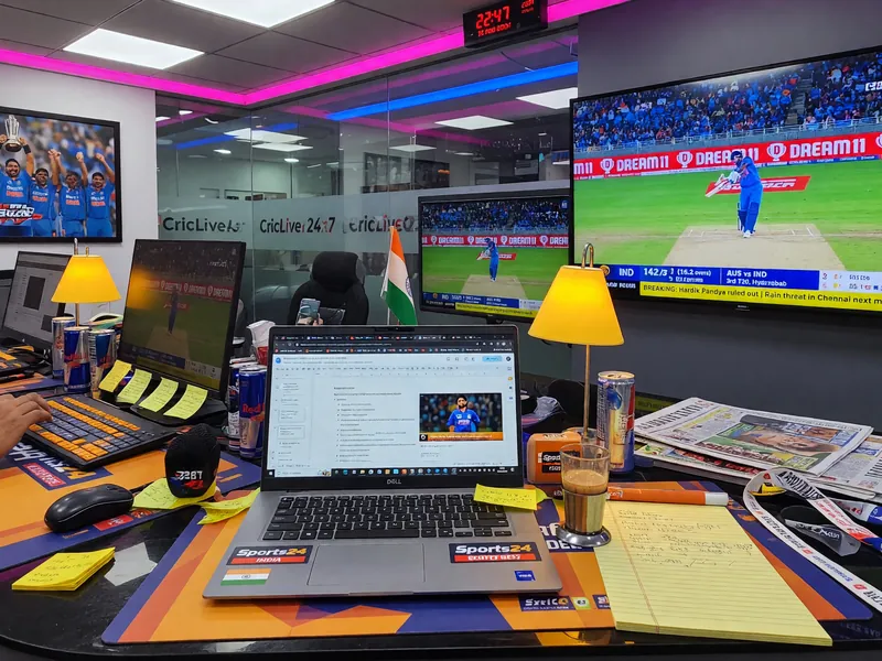 An editorial desk in front of a wall of monitors showing live match data, sports journalism setting